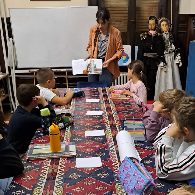 Voditeljka i deca za zajedničkim stolom tokom radionice u biblioteci